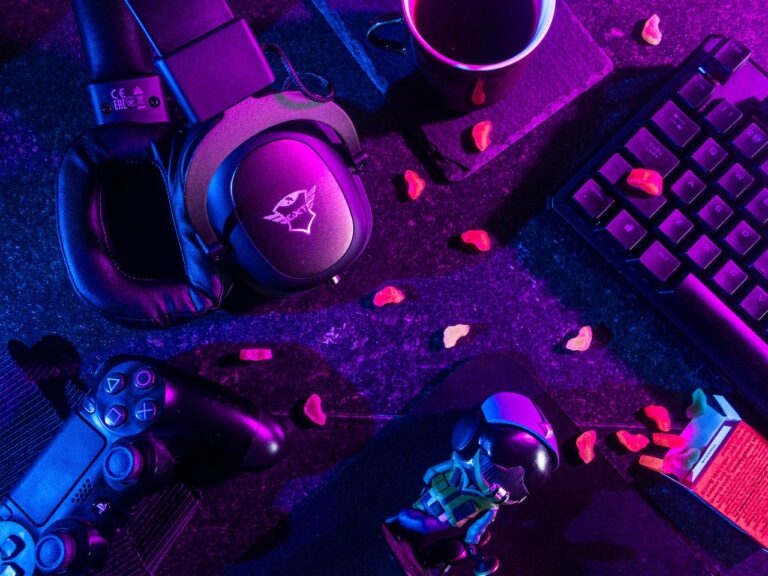 Das Bild zeigt eine Gaming-Szenerie bei künstlichem Licht, das einen violetten Farbton erzeugt. Zu sehen sind ein Headset auf der linken Seite, das auf einem Tisch liegt, neben einer Tasse auf einem Untersetzer. Rechts befindet sich eine beleuchtete Gaming-Tastatur mit einzelnen herzförmigen Konfetti-Stücken darauf verteilt. In der Mitte des Bildes ist ein Gaming-Controller zu erkennen und darunter eine Figur, die einen futuristischen Soldaten oder Charakter darstellen könnte.