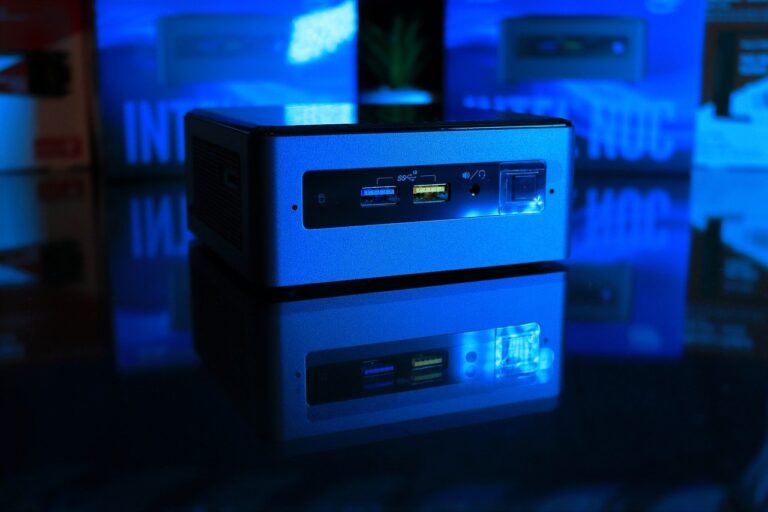 Das Bild zeigt einen einzelnen Mini-PC mit einem blauen Gehäuse, der auf einer reflektierenden Oberfläche steht, was zu einer deutlichen Spiegelung führt und den Eindruck erweckt, als ob zwei identische Geräte übereinander gestapelt wären. Der Mini-PC ist beleuchtet mit blauem Licht, welches die Szene in ein kühles technologisches Flair taucht. Auf der Vorderseite des Mini-PCs sind mehrere Anschlüsse sichtbar, darunter USB-Ports und ein Audio-Eingang, was auf eine vielseitige Konnektivität hinweist. Die Spiegelung und das blaue Licht verleihen dem Bild eine moderne und futuristische Atmosphäre.