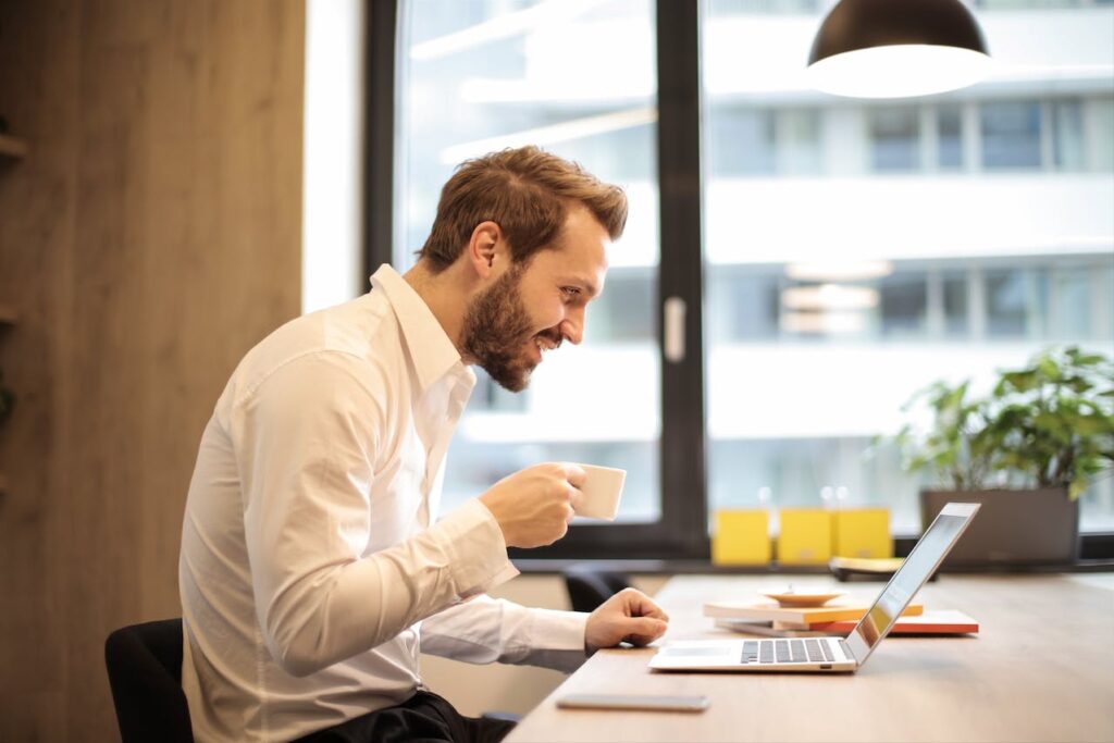 Das Bild zeigt einen Mann in einem gut beleuchteten Büro, der an einem modernen Schreibtisch vor einem Laptop sitzt. Er trägt ein weißes Hemd und scheint gerade eine Kaffeetasse zu halten, während er auf den Bildschirm seines Laptops blickt. Der Mann lächelt leicht, was darauf hindeutet, dass er möglicherweise eine positive Nachricht liest oder eine angenehme Arbeitserfahrung hat. Im Hintergrund sind große Fenster, die viel Tageslicht hereinlassen, und ein weiterer Arbeitsplatz zu sehen. Die Szene vermittelt eine professionelle und entspannte Arbeitsatmosphäre.