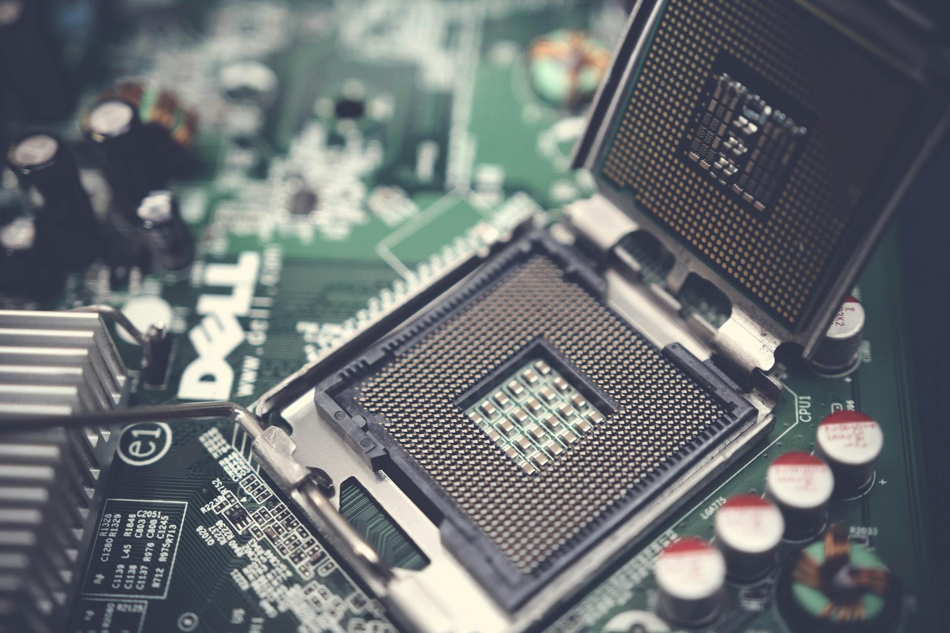 Das Bild zeigt einen offenen CPU-Sockel auf einer Dell-Mainboard-Platine. Der Sockel, ein integraler Bestandteil des Mainboards, ist bereit zur Aufnahme eines Mikroprozessors. Man erkennt die vielen kleinen, goldfarbenen Kontakte im Sockel, die eine präzise Verbindung zum Prozessor herstellen. Um den Sockel herum sind verschiedene elektronische Komponenten wie Kondensatoren, Widerstände und Spannungsregler zu sehen, die für das reibungslose Funktionieren des Mainboards wichtig sind. Der Fokus liegt auf dem Sockel, während die umgebenden Bauteile leicht unscharf im Hintergrund verschwimmen, was die zentrale Rolle des Sockels auf der Platine unterstreicht. Die Farbgebung ist kühl und technisch, mit einem starken Kontrast zwischen dem metallischen Grau des Sockels und dem grünen Mainboard.