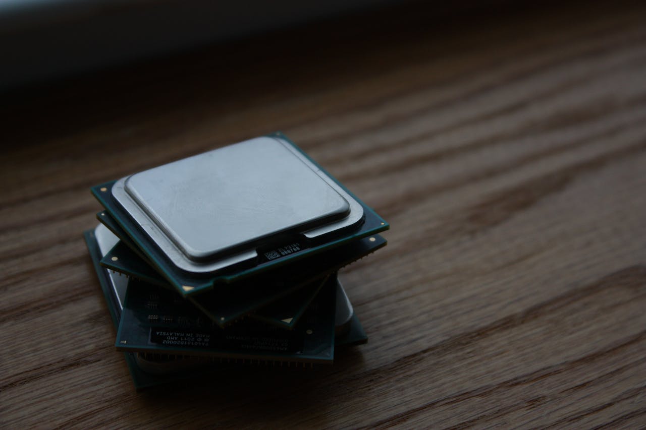 Das Bild zeigt einen Stapel von drei Prozessoren auf einer hölzernen Oberfläche. Die CPUs sind sichtbar gebraucht mit erkennbaren Abnutzungsspuren auf dem Metalldeckel, was auf eine frühere Nutzung hinweist. Die oberste CPU steht im Fokus, während die anderen beiden leicht unscharf sind, was eine Tiefenwirkung erzeugt. Das Foto ist in einem dunklen Raum mit natürlicher Beleuchtung aufgenommen, was zu einem weichen Schattenwurf und einer ruhigen Atmosphäre führt. Das Bild könnte die Evolution der Computerhardware oder die Thematik der technischen Aufrüstung symbolisieren.