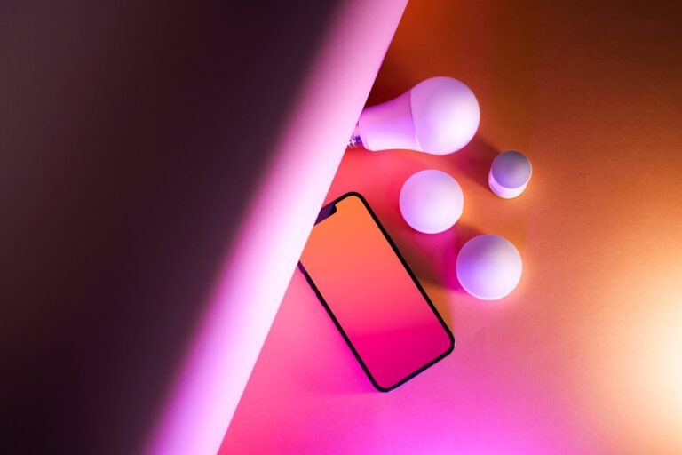 Ein modernes Smartphone, umgeben von intelligenten IoT-Glühbirnen, auf einer Oberfläche mit farbigem Verlauf, symbolisiert Vernetzung und Smart-Home-Technologie.