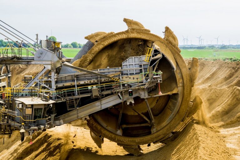 Ein riesiger Schaufelradbagger in Betrieb, der Sand in einer Tagebau-Mine fördert. Die gigantische Stahlkonstruktion ist mit mehreren Etagen, Treppen und Förderbändern ausgestattet. Im Hintergrund ist eine flache, landwirtschaftlich genutzte Landschaft mit mehreren Windkraftanlagen zu sehen. Die Szene vermittelt einen starken Kontrast zwischen schwerer Industriemaschinerie und erneuerbaren Energiequellen.