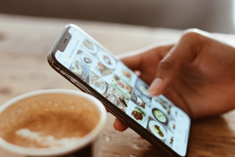 Ein Smartphone wird von einer Hand gehalten, während darauf Bilder von Lebensmitteln auf einer Social Media Plattform angezeigt werden. Im Vordergrund ist eine Tasse Kaffee zu sehen.