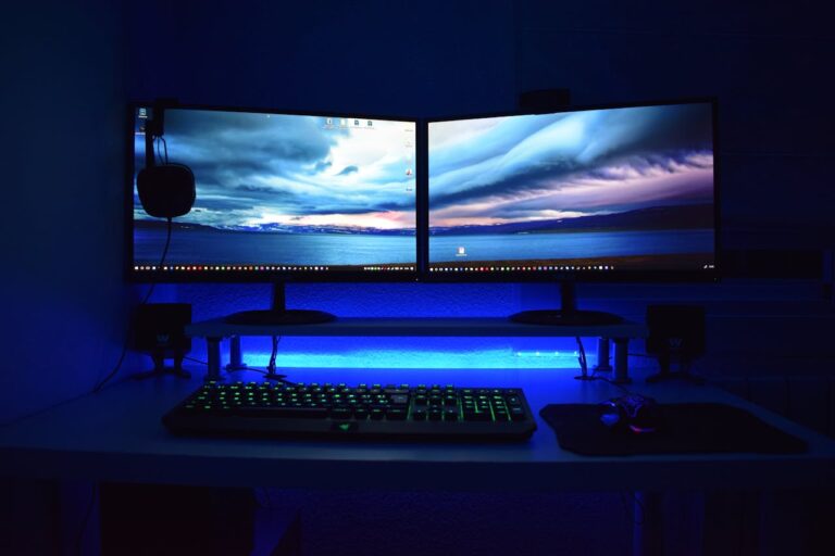 Ein modern eingerichtetes Gaming-Setup in einem dunklen Raum, beleuchtet von blauem LED-Licht. Es besteht aus zwei nebeneinander stehenden Monitoren, einer beleuchteten Gaming-Tastatur und einer Gaming-Maus auf einem Mauspad. Ein Headset hängt an der linken Seite des linken Monitors. Die Monitore zeigen einen Desktop-Hintergrund mit einem Bild eines bewölkten Himmels über einem See.