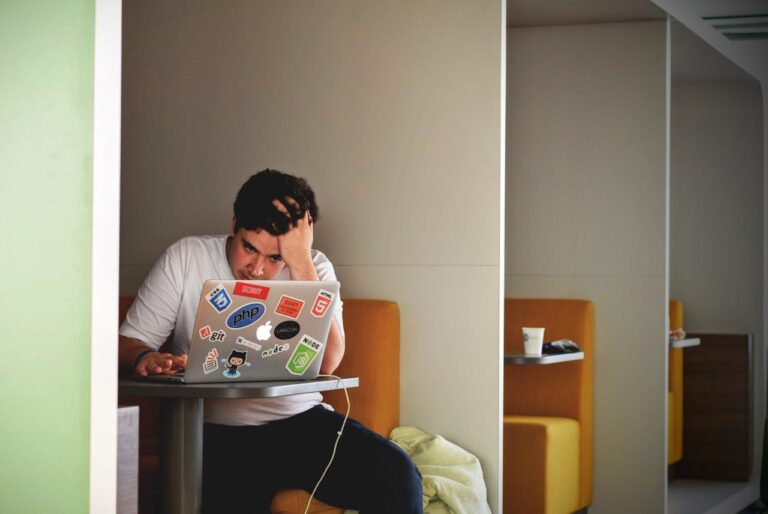 Ein Mann sitzt in einer modernen Sitznische und arbeitet konzentriert an seinem Laptop. Er stützt seinen Kopf auf eine Hand, was auf Frustration oder Nachdenklichkeit hindeutet. Der Laptop ist mit verschiedenen Programmier- und IT-bezogenen Stickern beklebt. Die Szene wirkt ruhig und isoliert, mit einem minimalistischen Hintergrund und warmen Farben.