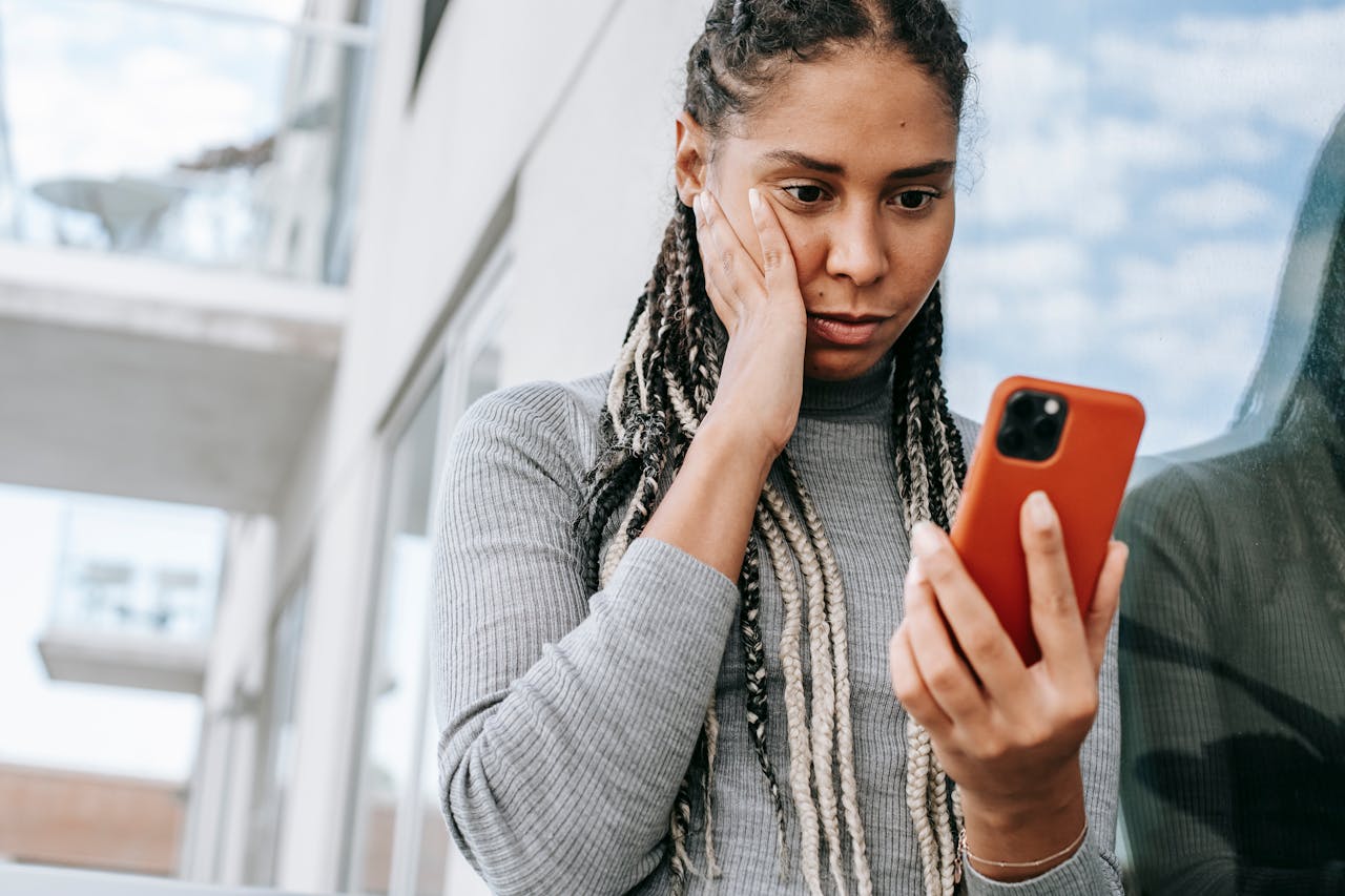 Eine besorgte Frau mit langen, geflochtenen Haaren hält ein Smartphone in der Hand und blickt nachdenklich auf den Bildschirm. Sie steht draußen vor einem modernen Gebäude mit Glasfassade.