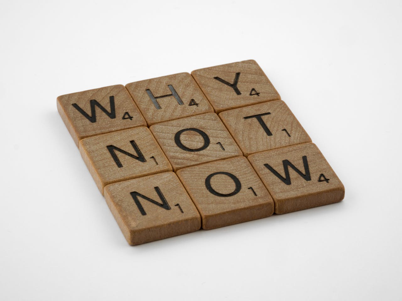 Scrabble-Buchstaben formen die Worte 'WHY NOT NOW' auf einem hellen Hintergrund, was symbolisch für Entschlossenheit oder die Aufforderung zu sofortigem Handeln steht.