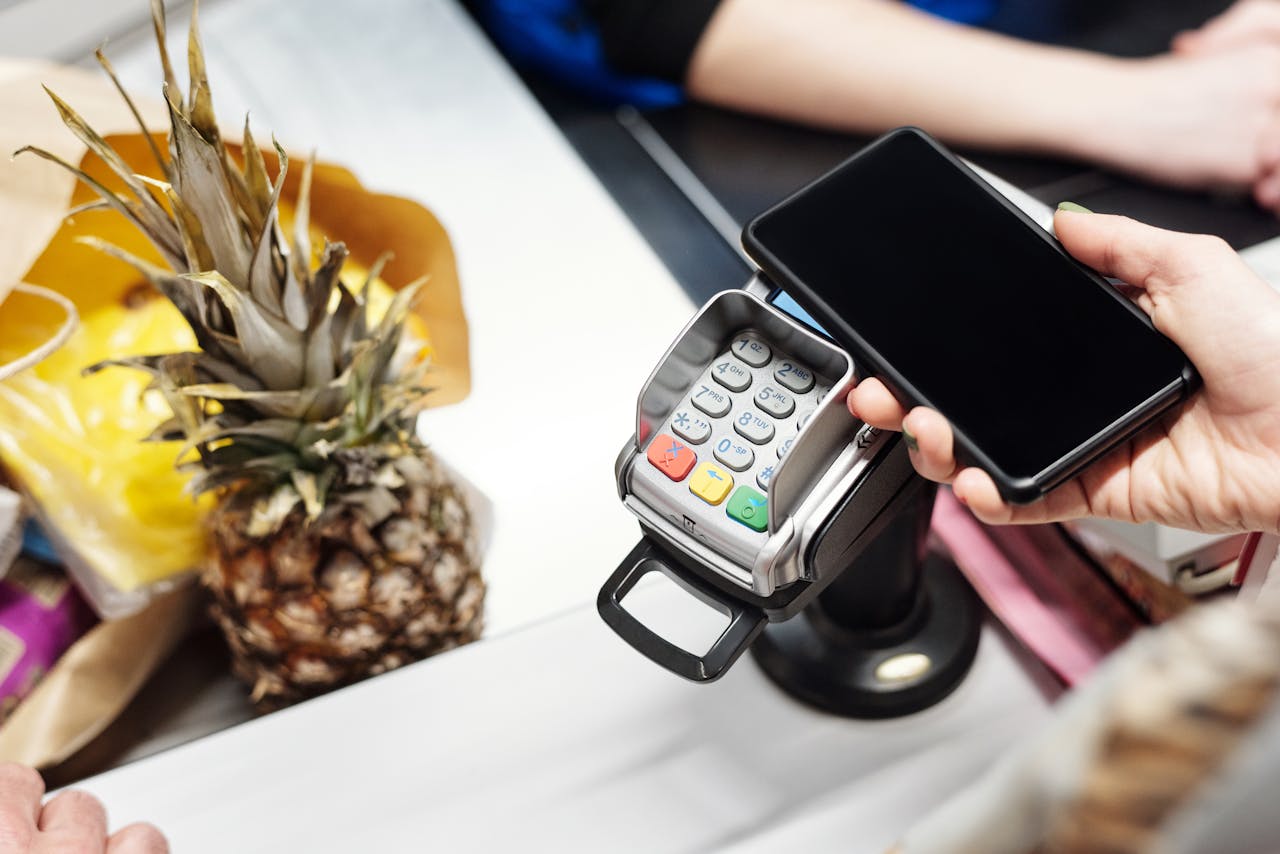 Eine Person hält ein Smartphone an ein Zahlungsterminal, um eine NFC-Zahlung im Geschäft durchzuführen. Im Hintergrund sind Einkäufe wie eine Ananas zu sehen.