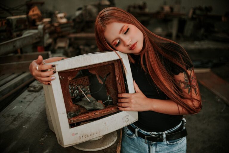 Eine junge Frau mit roten Haaren lehnt ihren Kopf sanft an einen alten, verrosteten Röhrenmonitor mit beschädigtem Inneren. Der Monitor steht auf einem Tisch in einer industriellen Umgebung, die von Maschinen und Werkzeugen umgeben ist. Die Szene vermittelt einen nostalgischen und nachdenklichen Eindruck.