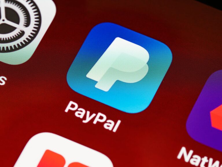 Ein Nahaufnahmefoto des PayPal-Logos auf einem Smartphone-Bildschirm, angezeigt neben anderen App-Symbolen auf einem roten Hintergrund.