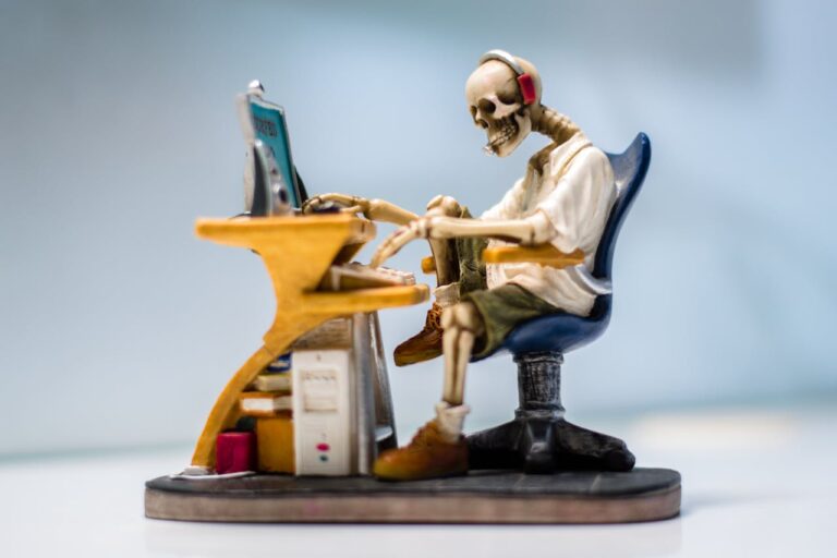 Eine kleine Figur eines Skeletts mit Kopfhörern sitzt an einem Schreibtisch vor einem Computer, was eine humorvolle Darstellung langer Computer-Sitzungen symbolisiert.