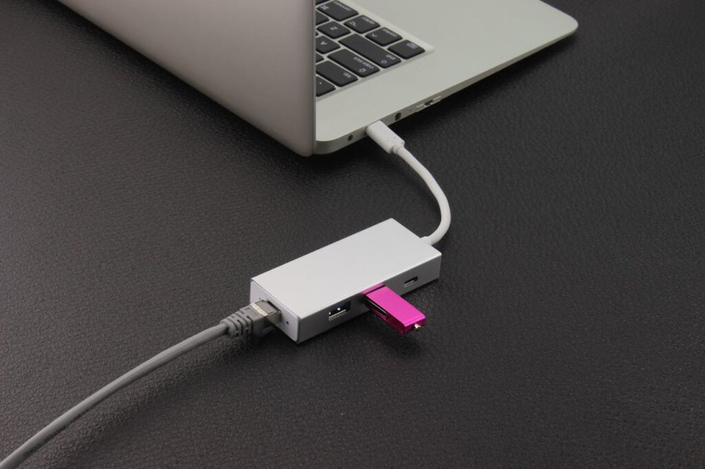 USB-Hub mit mehreren angeschlossenen Geräten, darunter ein pinkfarbener USB-Stick und ein Netzwerkkabel, verbunden mit einem silbernen Laptop auf schwarzer Oberfläche.